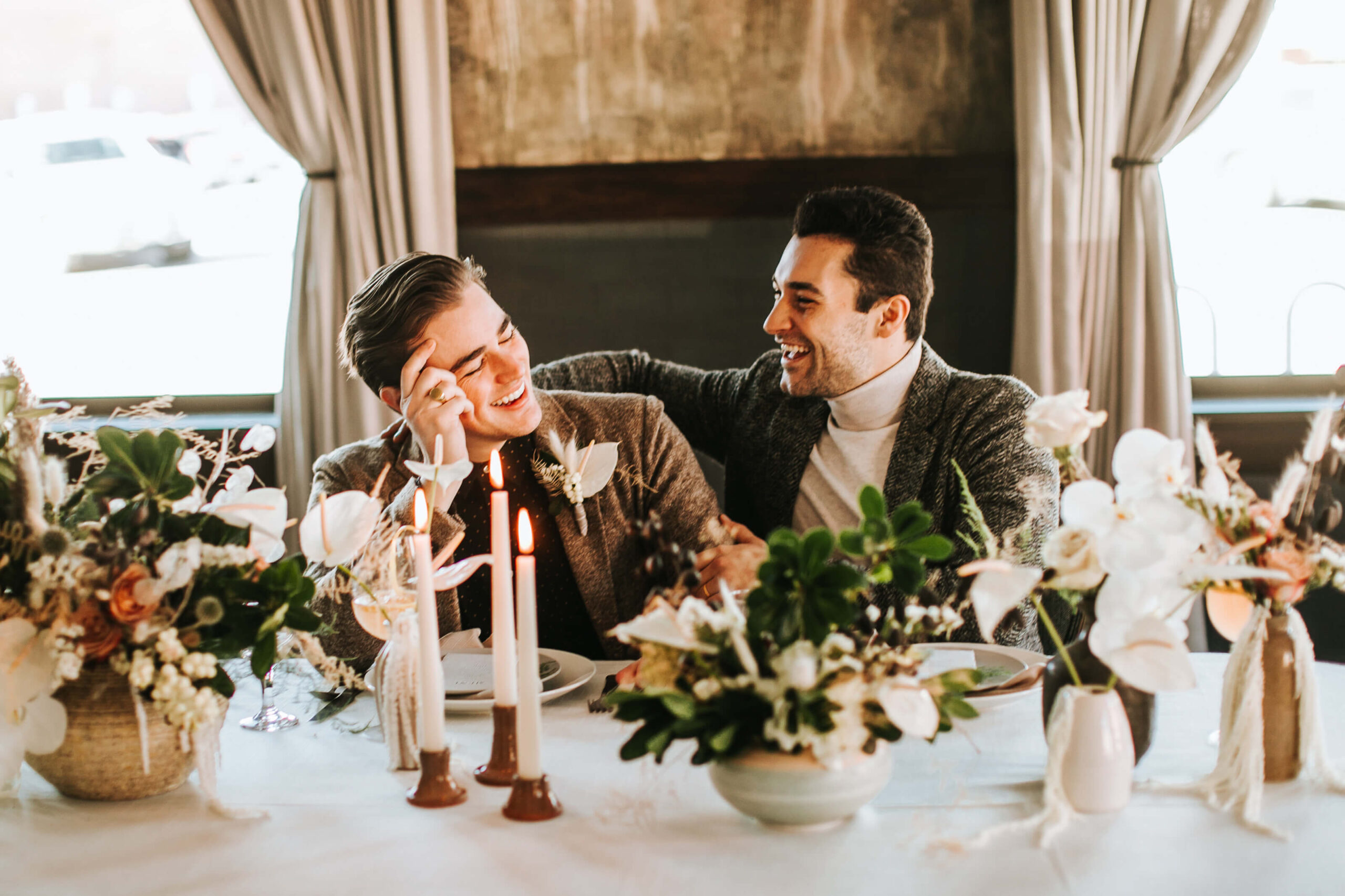 Same sex couple on wedding day, laughing at dinner table.